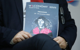 Uczestnik panelu dyskusyjnego trzyma na kolanach teczkę z napisem Welconomy Forum i podobizną Mikołaja Kopernika