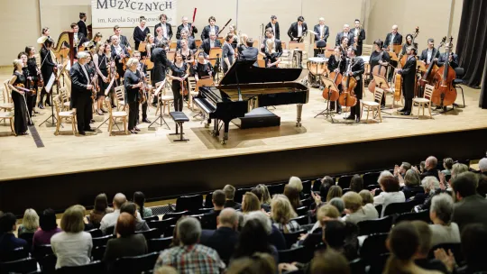 muzycy stoją na scenie, na środku fortepian, w tle napis zespół szkół muzycznych, przed sceną klaszcząca widownia