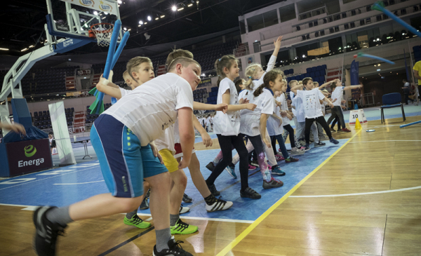Dzieci uczestniczą w zawodach w hali sportowej, stoją na linii i rzucają piłkami