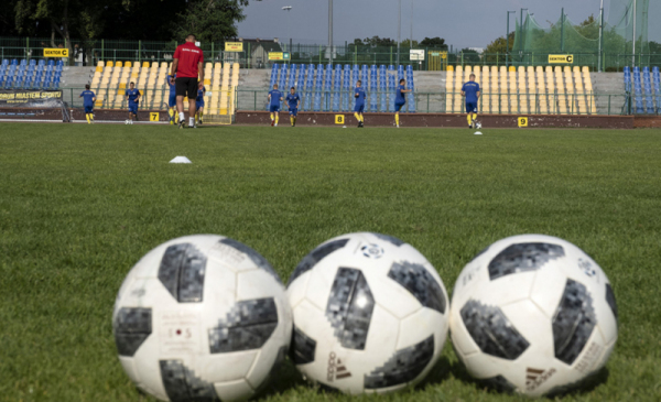Na zdjęciu: trzy piłki do piłki nożnej leżą na zielonej murawie stadionu, w tle trybuny pomalowane w żółto-niebieskie barwy klubu Elana