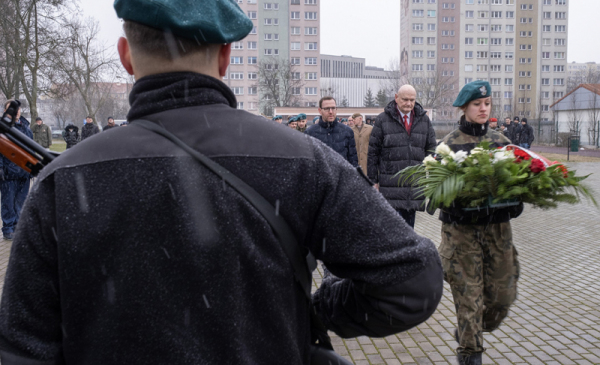 Na zdjęciu widać plecy żołnierza stojcego na warcie obok młoda dziewczyna w mundurze trzyma wieniec kwiatów
