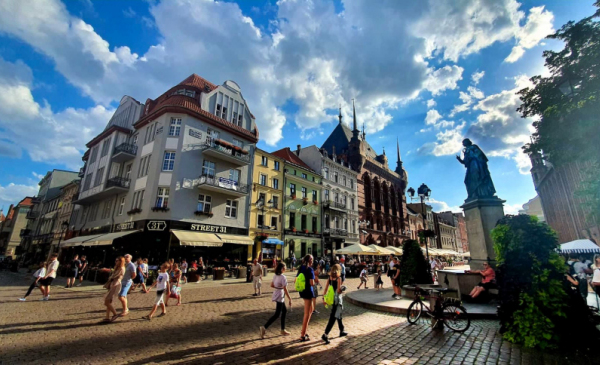 Na zdjeciu: turyści idą przez Rynek Staromiejski, obok pomnik Kopernika, w tle błękitne niebo z białymi obłokami