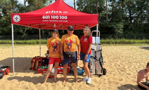 Na zdjęciu: troje ratowników wodnych stoi na plaży, za nimi widać czerwony namiot z białymi napisami z numerem telefonu do WOPR