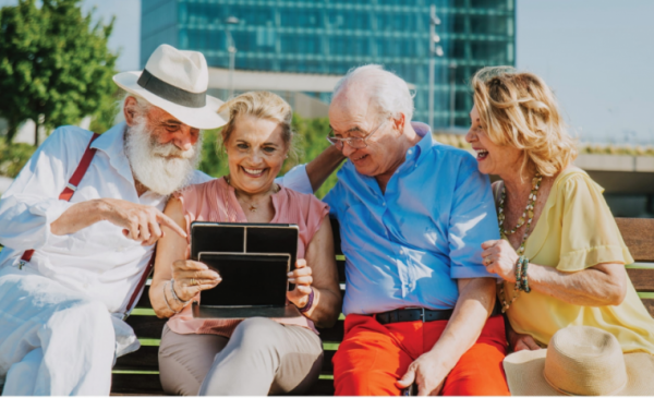 Zdjęcie przedstawia uśmiechniętych seniorów, którzy oglądają coś na tablecie. W tle widnieje budynek ZUS