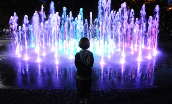 Na zdjęciu: kolorowe podświetlenia fontanny Cosmopolis nocą, fioletowo-błekitne, na ich tle ciemna sylwetka zapatrzonego dziecka