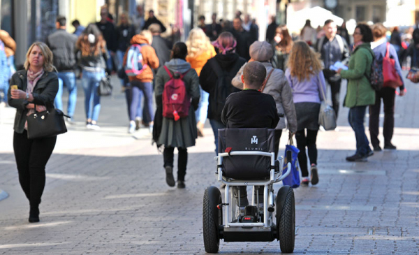 Na zdjęciu: osoba na wózku jedzie chodnikiem, wokół spacerują ludzie