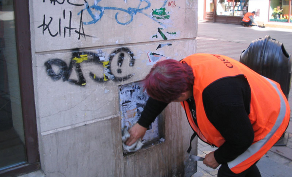 Osoba w pomarańczowej kamizelce myjącą ściane pomazaną graffiti