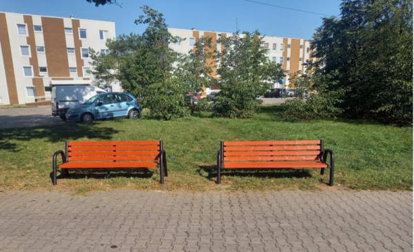 Na zdjęciu dwie ławeczki na skwerze, w tle bloki mieszkalne