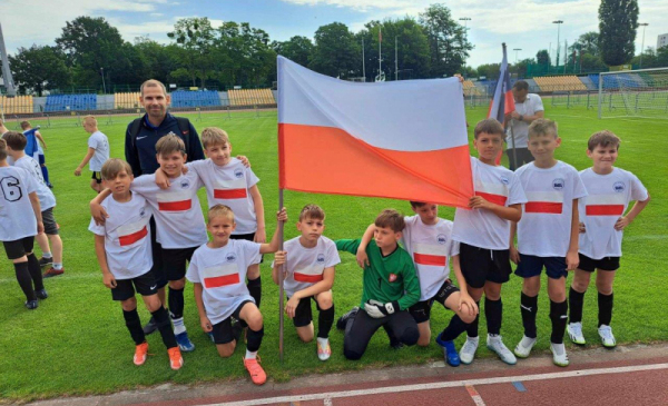 Na zdjęciu: dzieci na zielonej murawie piłkarskiej trzymają flagę Polski
