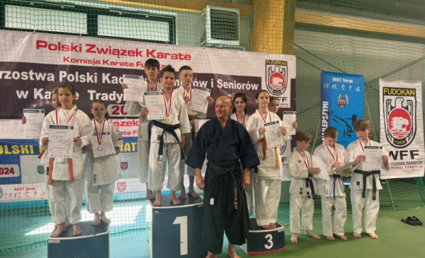 Na zdjęciu: dzieci w strojach karate stoją na podium, przed nimi trener w czarnej judoce