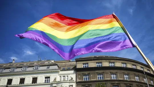 Tęczowa flaga powiewa na tle niebieskoiego nieba.