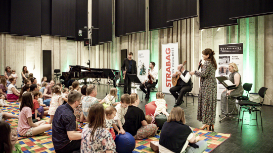 dzieci i rodzice siedzą na kolorowym dywanie w sali prób orkiestry, przed nimi muzycy i prowadząca z mikrofonem, która się śmieje