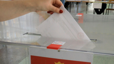 Na zdjęcli: dłoń o czerwonych paznokciach trzyma kartę do głosowania i wrzuca ją do urny wyborczej, na której jest godło kraju