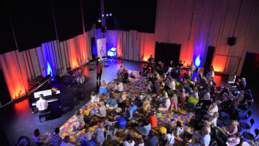 dzieci i rodzice siedzący na kolorowym dywanie i poduchach w sali prób orkiestry, przed nimi muzycy