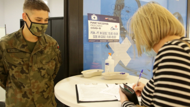 Na zdjęciu kobieta wypełnia formularz szczepienny, obok stoi żołnierz WOT
