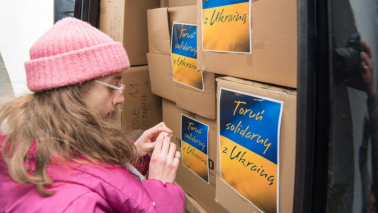 Na zdj: dziewczyna w różowej kurtce i czapce przykleja plakat z flagą Ukrainy i napisem Solidarni z Ukrainą na kartony z artykułami pierwszej potrzeby