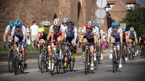 Na zdjęciu: kolarki jadą na rowerach w wyścigu, obok mury gotyckie Torunia