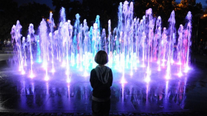 Na zdjęciu dziecko od tyłu wpatruje się w podświetloną na niebiesko i fioletowo fontannę 