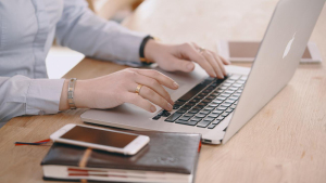 Na zdjęciu widać dłonie kobiety, pracującej na laptopie, obok leżą notes i telefon