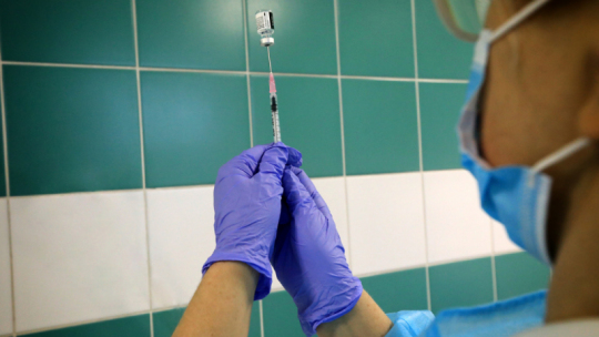 Na zdjęciu: widać dłonie pielęgniarki w fioletowych rękawiczkach trzymające strzykawkę z dawką szczepionki