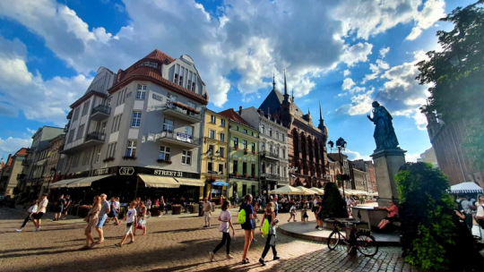 Na zdjeciu: turyści idą przez Rynek Staromiejski, obok pomnik Kopernika, w tle błękitne niebo z białymi obłokami