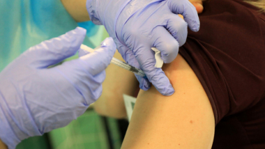 Na zdjęciu widać dłonie pielęgniarki w fioletowych rękawiczkach podczas wykonywaniu zabiegu szczepienia - igła wkłuwana jest w ramię pacjenta