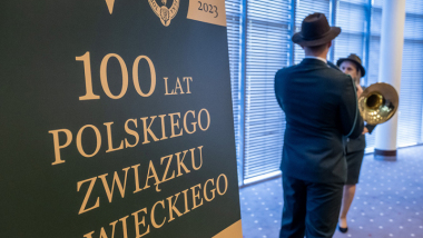 Na zdjęciu: obok  rollupu informującego o 100-leciu Polskiego Związku Łowieckiego stoi Łowczy w tradycyjnym stroju w kolorze ciemnozielonym i gra na trąbce