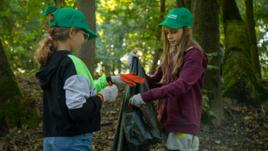 Na zdjęciu: dwie dziewczynki w zielonych czapeczkach z daszkiem i rękawicach ochronnych trzymają worek ze śmieciami