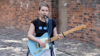 Na zdjęciu: młoda dziewczyna śpiewa do mikrofony, gra na niebieskiej gitarze elektrycznej, w tle ceglany mur