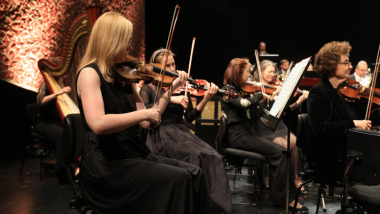 Na zdjęciu: gra orkiestra, na pierwszym planie skrzypaczka o jasnych włosach