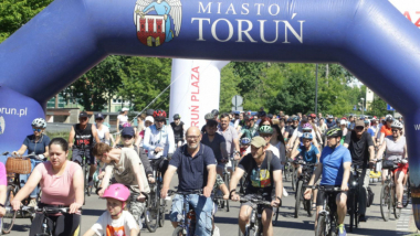 Na zdjęciu: ludzie jadą na rowerach, nad nimi duży napis Miasto Toruń i herb Torunia