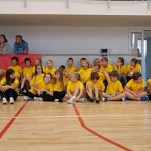 Na zdjęciu: w sali sportowej siedzą na podłodze dzieci w żółtych koszulkach