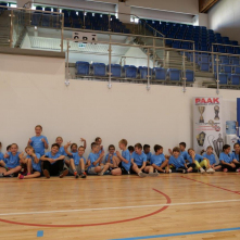Na zdjęciu: w sali sportowej siedzą na podłodze dzieci w niebieskich koszulkach