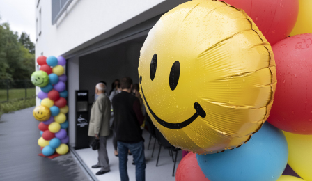 Na zdjęciu widac żółty balon z namalowaną uśmiechniętą buźką, w tle goście uroczystości otwarcia żłobka stoją przy budynku