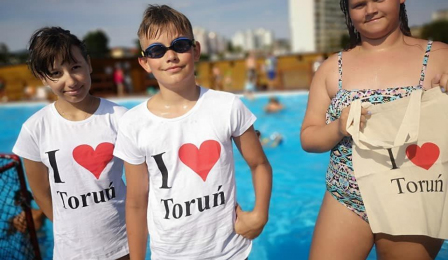 Na zdjęciu stoją przy basenie letnim dziewczynki ubrane w koszulki z napisem "I love Toruń"