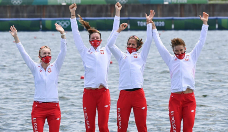 Zawodniczki czwórki podwójnej w białoczerwonych dresach i maseczkach machają przed dekoracją olimpijską 